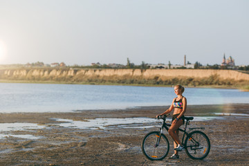 Fototapeta premium A strong blonde woman in a colorful suit sits on a bicycle in a desert area near the water and looks at the sun. Fitness concept. Blue sky background