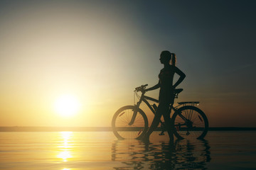 Fototapeta na wymiar A strong blonde woman in a colorful suit stands near the bicycle in the water at sunset on a warm summer day. Fitness concept. Sky background