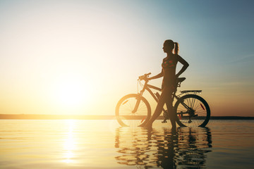 A strong blonde woman in a colorful suit stands near the bicycle in the water at sunset on a warm summer day. Fitness concept. Sky background