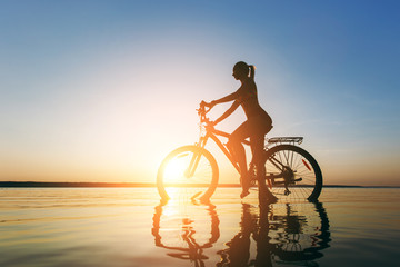 A strong blonde woman in a colorful suit sits on the bicycle in the water at sunset on a warm summer day. Fitness concept. Sky background