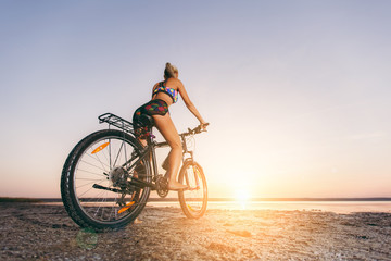 Fototapeta na wymiar A strong blonde woman in a multicolored suit sits on a bicycle in a desert area near the water and looks at the sun. Fitness concept. Back view