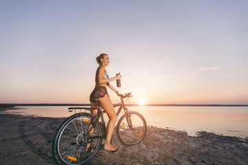 A strong blonde woman in a colorful suit sits on the bicycle, holds black bottle with water in a desert area near the water. Fitness concept. Blue sky background