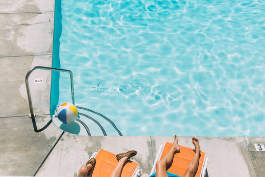 Legs Of Man And Woman Lounging On Orange Lawn Chairs By Swimming Pool