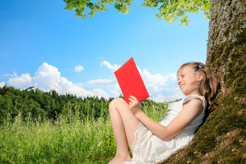 young girl sit next to a tree and smile into the summer sun. she plays with electronic devices, red apple, flowers. sky is blue and the grass is green.