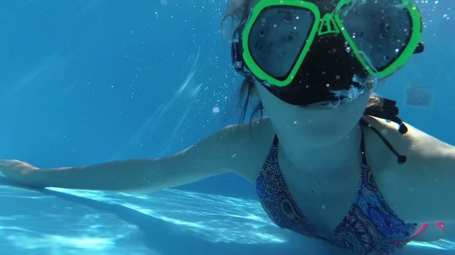 Happy girl in a mask under the water in the swimming pool looks into the camera, bubbles, sun rays