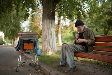 Homeless man sitting on bench lightning cigarette. Old man with no place of living smoke in the city park.