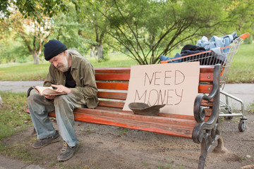 View of tramp in old dirty clothes sitting on bench reading a book. On bench old homeless beggar and board with sign need money.