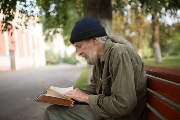 Fototapeta premium Side view of old homeless man with long grey hair reading a book. Tramp sitting on the bench in the street,