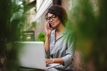 Beautiful African American girl in glasses sitting on bench and talking on cellphone while dreamily looking in laptop. Young lady with dark curly hair sitting at table on street and working on laptop