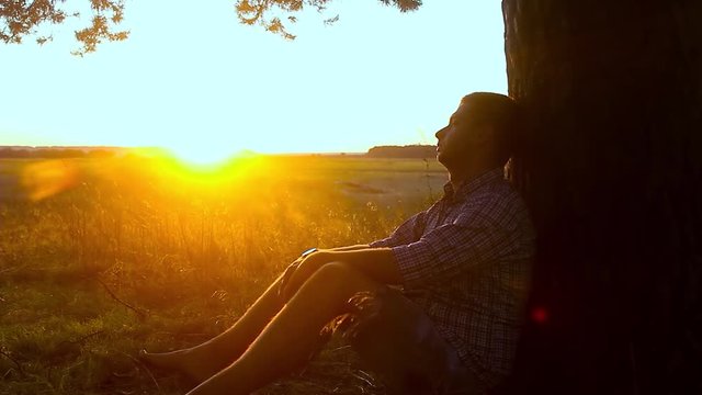 Human sitting under tree. Man repose on grass in nature. Outdoors - outside. Young man meditating in half lotus - beautiful sunset as a background.