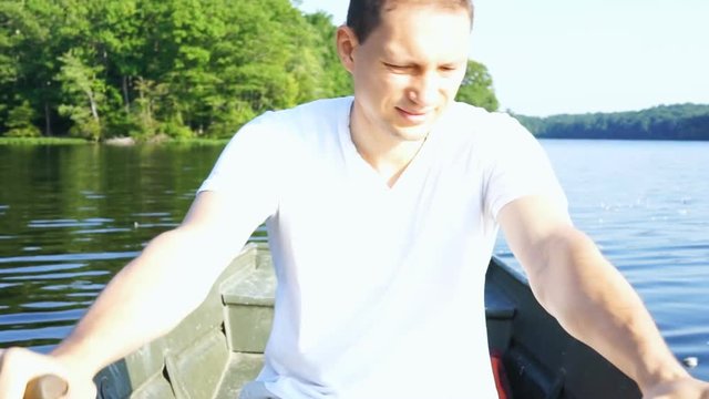 Happy smiling young man rowing boat on lake in Virginia during summer in white shirt, looking back, steering