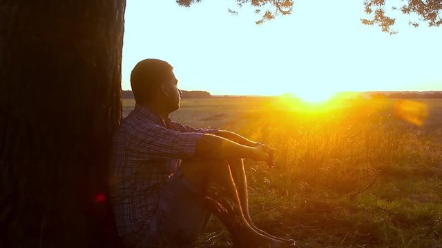 Human sitting under tree. Man repose on grass in nature. Outdoors - outside. Young man meditating in half lotus - beautiful sunset as a background.