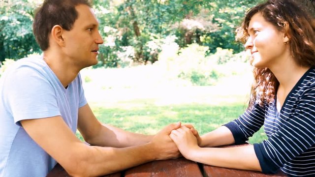 Young Couple Sitting, Talking, Holding Hands At Picnic Table In Outdoor Park Making Up With Man Kissing Hand At The End