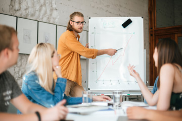 Fototapeta premium Portrait of young man standing near board and speaking with colleagues in office . Group of young people sitting at the table on business meeting and discussing diagram