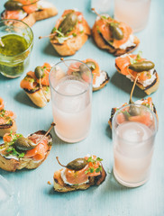 Crostini with smoked salmon, pesto sauce, watercress and capers and pink grapefruit cocktails in glasses over light blue background, selective focus. Party, catering or fingerfood concept