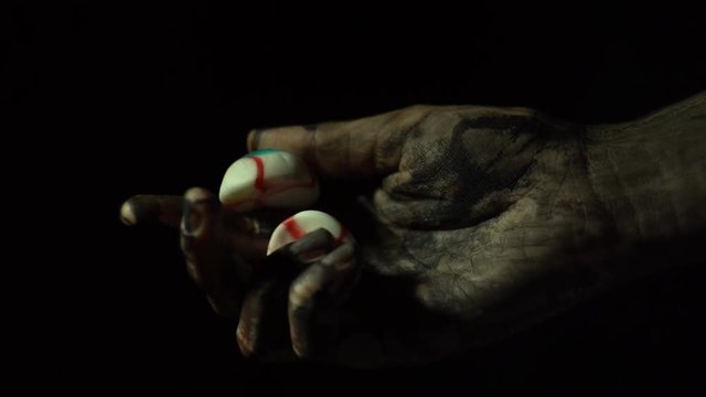A close-up dirty zombie hand holds and rotates two eyes on a black background in a dark room.