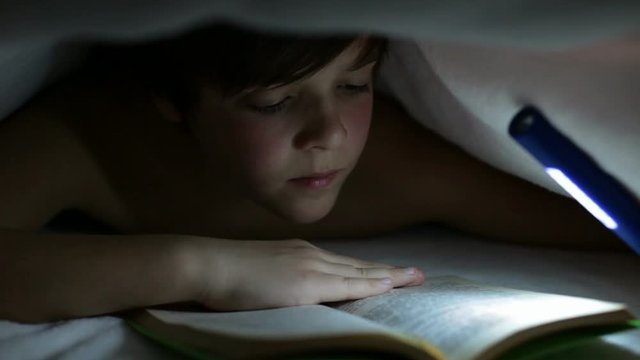 Young boy reading a captivating book at night - lighting with a flashlight under the blanket