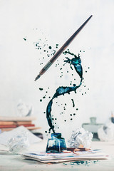 Spilled ink flying above inkwell in a spiraling splash with tiny drops and flying pen on a light background. Still life with writer workplace. Creative writing concept. - obrazy, fototapety, plakaty