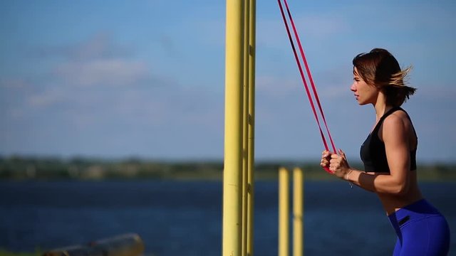 Brunette in blue leggings on the river Bank performs exercises on the triceps with a rubber band.
