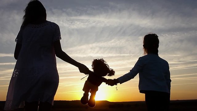 Silhouette of mom and daughter at sunset, holding toy doll and staring into the distance. Silhouette of girl play with dolls