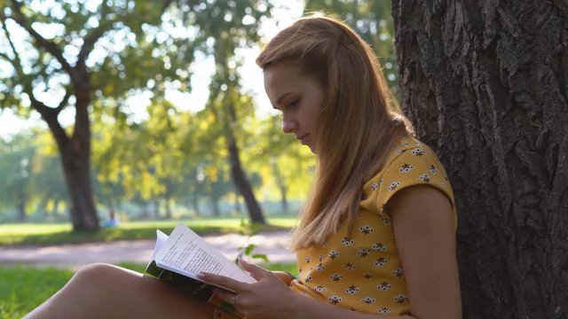 A young blond girl in a warm sunny day at a picnic. She is dressed in a bright yellow dress. green grass . holds in his hand a book and reads. rest in the park. the woman is sitting near a big tree