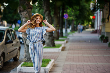 Portrait of a very attractive young woman in striped overall posing with her hat on a pavement in a town with trees in a background.