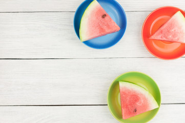 Three slices of fresh ripe watermelon on green red and blue ceramic plates on old wooden rustic white table