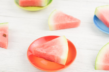 Three slices of fresh ripe watermelon on green red and blue ceramic plates and scattered slices on old wooden rustic white table