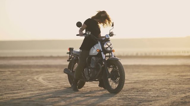 Attractive young woman motorcyclist makes a burnout on a motorcycle and ride away. Sunset or sunrise shot at desert. Female biker. Slow motion