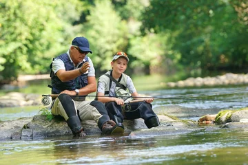 Zelfklevend Fotobehang Father and son fly-fishing in river, sitting on rocks © goodluz