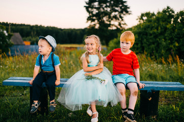 Fototapeta premium Group of little emotional kids sitting on bench outdoor in countryside. Girl in dress between two guys. Difficult relations. Youth jealousy. Funny loving triangle. Joy, sorrow, hurt and offence.
