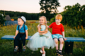 Group of little emotional kids sitting on bench outdoor in countryside. Girl in dress between two guys. Difficult relations. Youth jealousy. Funny loving triangle. Joy, sorrow, hurt and offence.