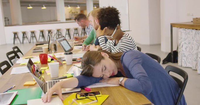 Young exhausted female employee lying on table and sleeping on background of working colleagues.