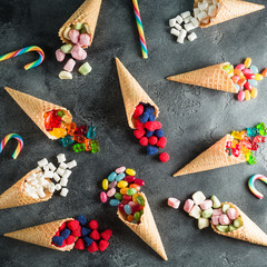 Pattern with colorful bright assorted candy in waffle cones on dark background. Flat lay, top view