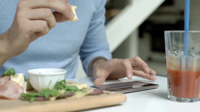 Man in blue sweater eating cheese while texting messages on smartphone
