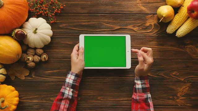 White tablet computer with green screen in horizontal position on the wooden table with autumn fruits and vegetables. Top view. Female finger scrolling pages. Fall. Chroma key
