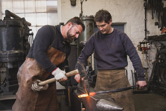 Two blacksmiths hammer a piece of metal on an anvil in a workshop.