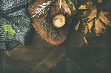 Autumn or Fall motning coffee concept. Flat-lay of arm knitted woolen grey sweater, wooden tray, mug of coffee and yellow fallen leaves over dark rustic wooden table background, top view, copy space.