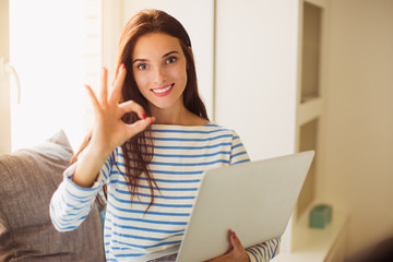 Portrait of young bewitching smiling woman who sitting on the sofa in the room and keeping laptop and showing sign 