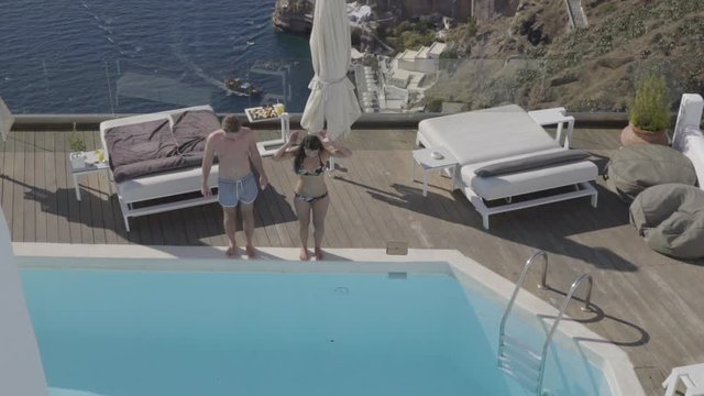 4k travel video slow motion couple jumping in swimming pool of luxury hotel terrace high above sae on volcanic island of santorini