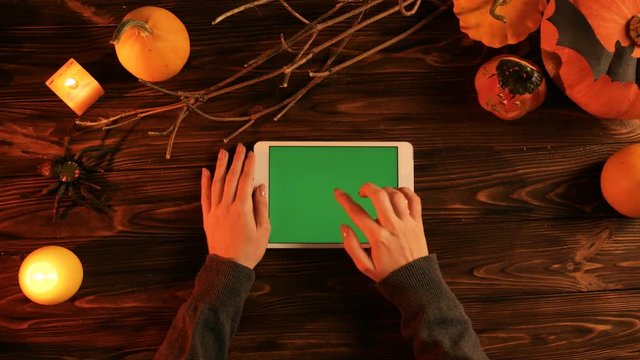 Halloween top view concept, Woman using white tablet computer with green screen on the wooden table. Flat lay with halloween decor. Pumpkins with candles in the dark. Chroma key