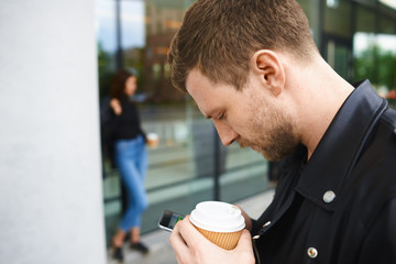 Outdoor picture of handsome young beaded male holding takeaway coffee in disposable papercup and typing text message using mobile phone while his stylish girlfriend standing on street in background