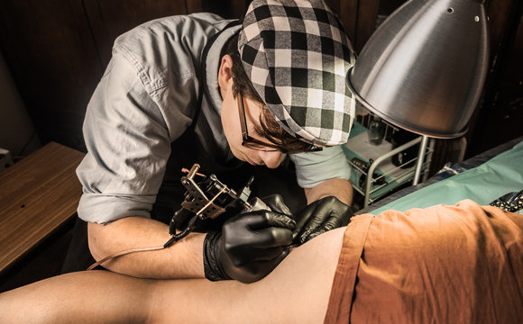 Young tattoo artist in vintage outfit inking female legs/look from above
