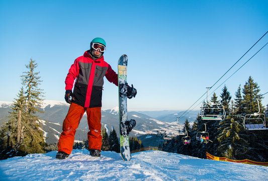 Full length shot of a snowboarder standing with his snowboard on the top of the mountain after riding at winter ski resort in the evening. Mountains, forests, ski lift and blue sky on the background