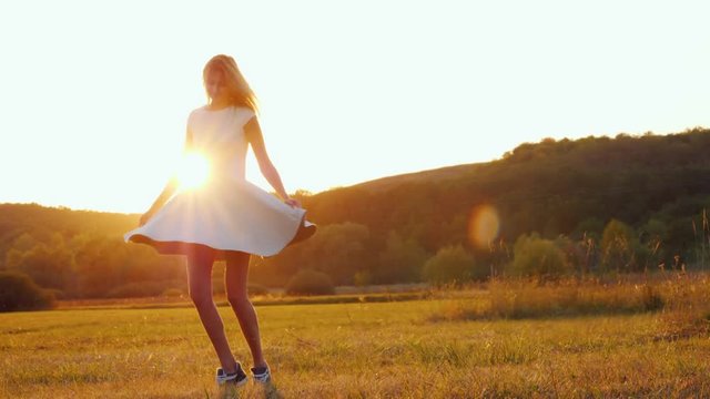 Young woman in a light dress whirls on a field in the rays of a sunset
