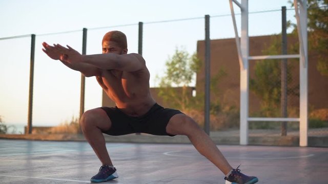 bearded blond-haired metis guy with naked torso squatting and stretching his body at sunrise time on basketball playground slomo 