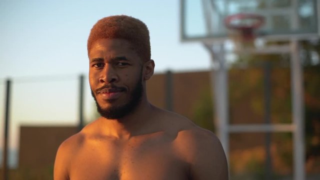 closeup slow motion at sunrise muscular guy with stylish haircut and naked torso looking at camera smiling standing on basketball court background in defocus 