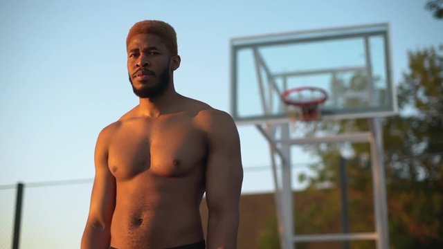 closeup slow motion at sunrise muscular guy with dyed hair and naked torso looking at camera smiling standing on basketball court background in defocus 