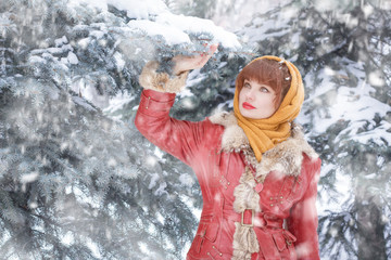 Fototapeta na wymiar Beautiful Slavic woman, in winter, in a headscarf on her head, looks out into the distance, against the background of coniferous trees. Russian winter, Big red lips. Snow falls