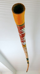 Unter einer Decke hängendes gelblich und braun bemaltes Didgeridoo, von unten gesehen